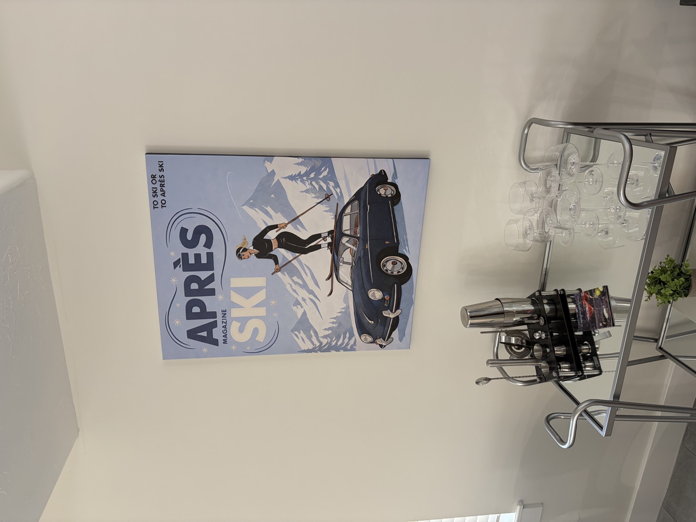 An Après Ski magazine-cover print, a skier standing on the roof of a navy Porsche 356 in the snow, hung above a chrome bar cart with a cocktail set and glasses.