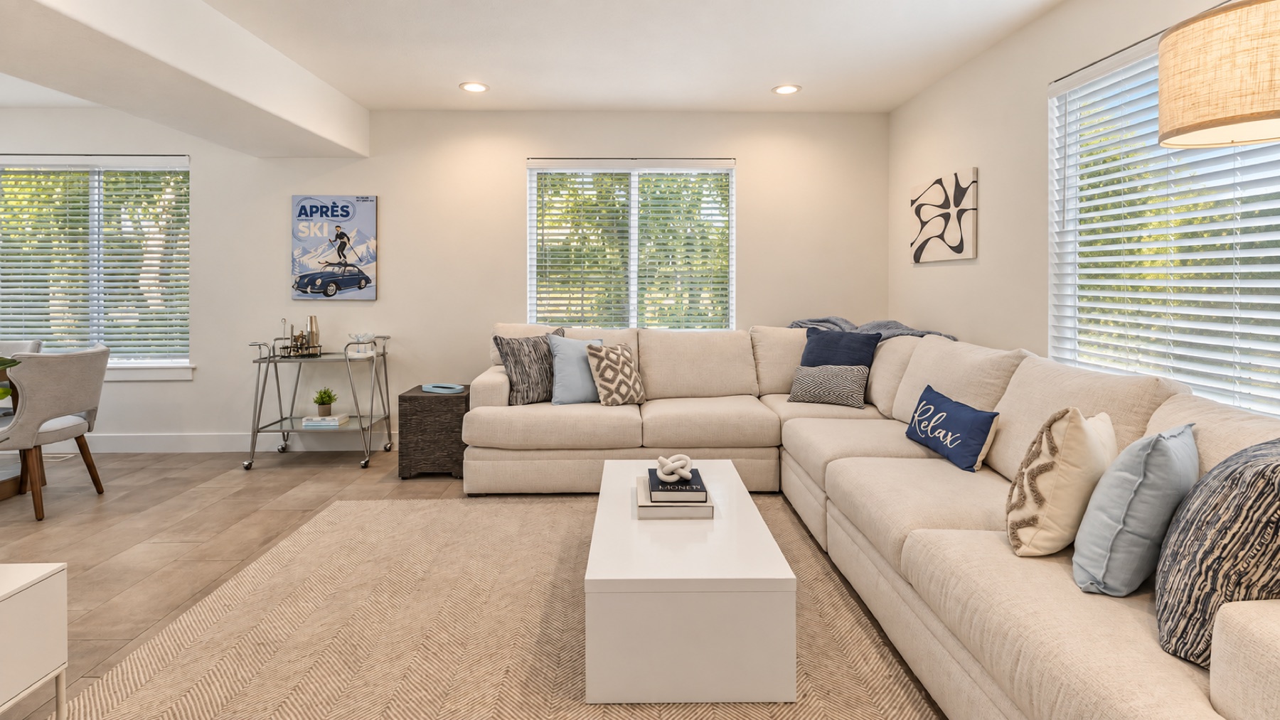 The room from the far corner: the Après Ski print over the bar cart, three windows, the long side of the sectional.