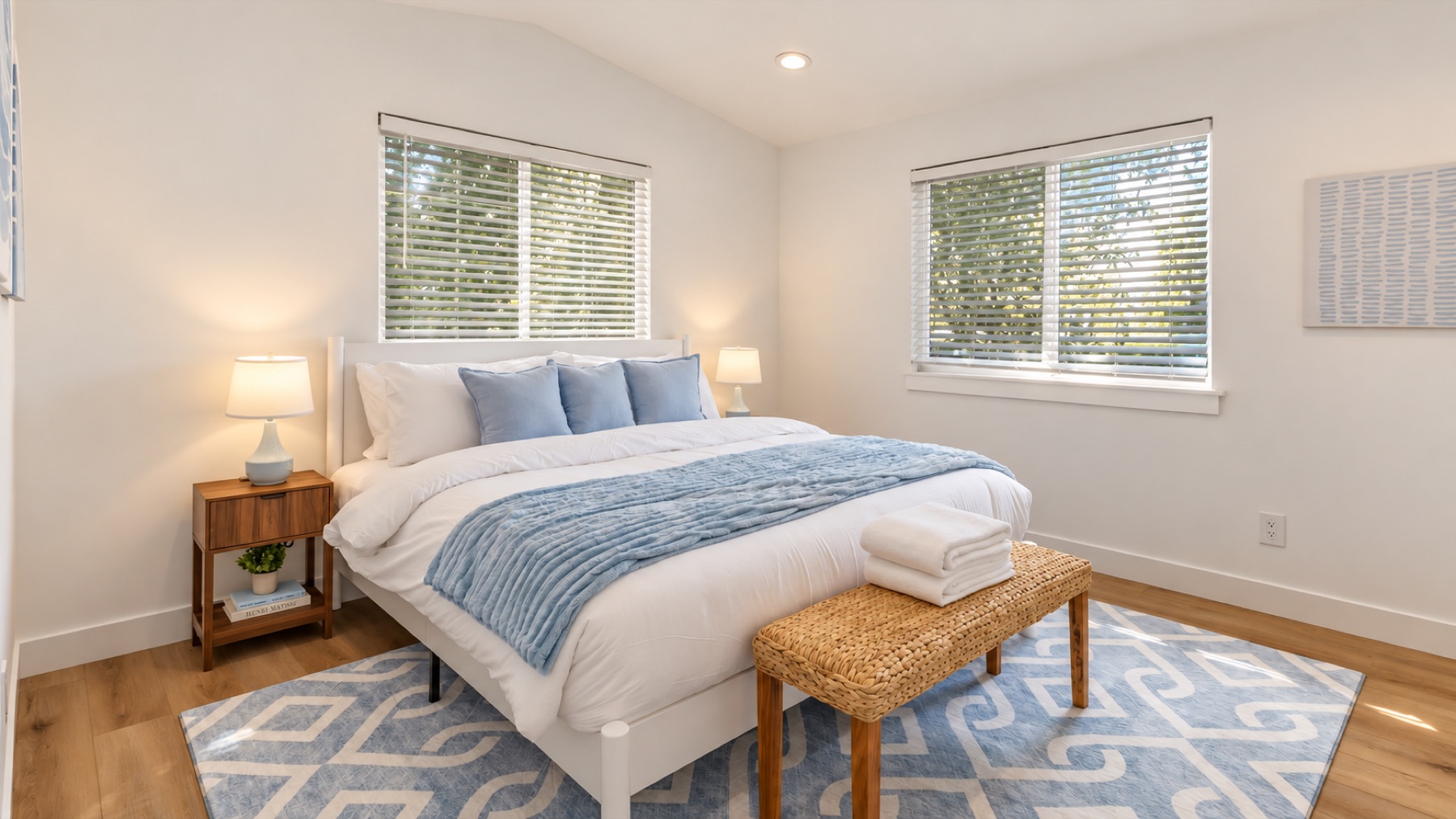 The primary bedroom, in blues: a white bed with a blue throw, two windows, a rattan bench at the foot, and its own bath.
