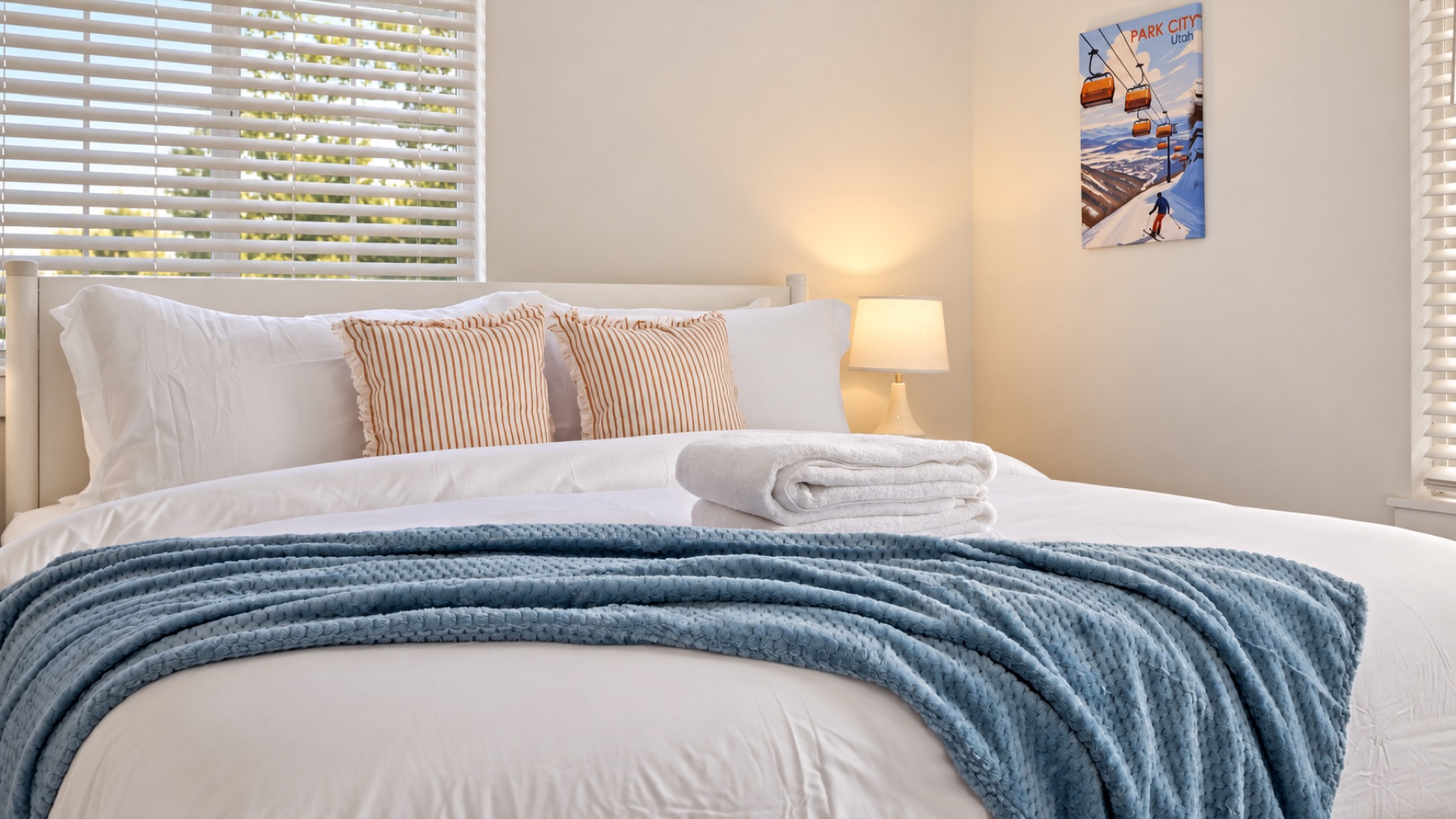 Bedroom two made up in the morning: tan pillows, a blue throw, fresh towels folded at the foot. It shares the hall bath with bedroom three.