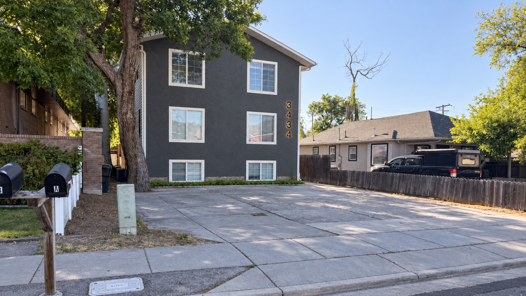 The building from the street: a charcoal two-storey house under a big tree, with a mailbox at the curb and off-street parking in front.