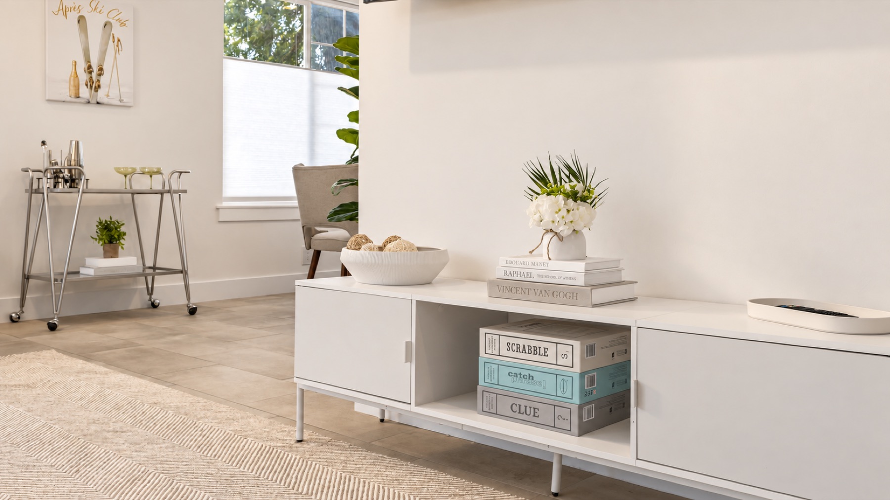 The console under the print, stacked with board games, and the chrome bar cart beside it.