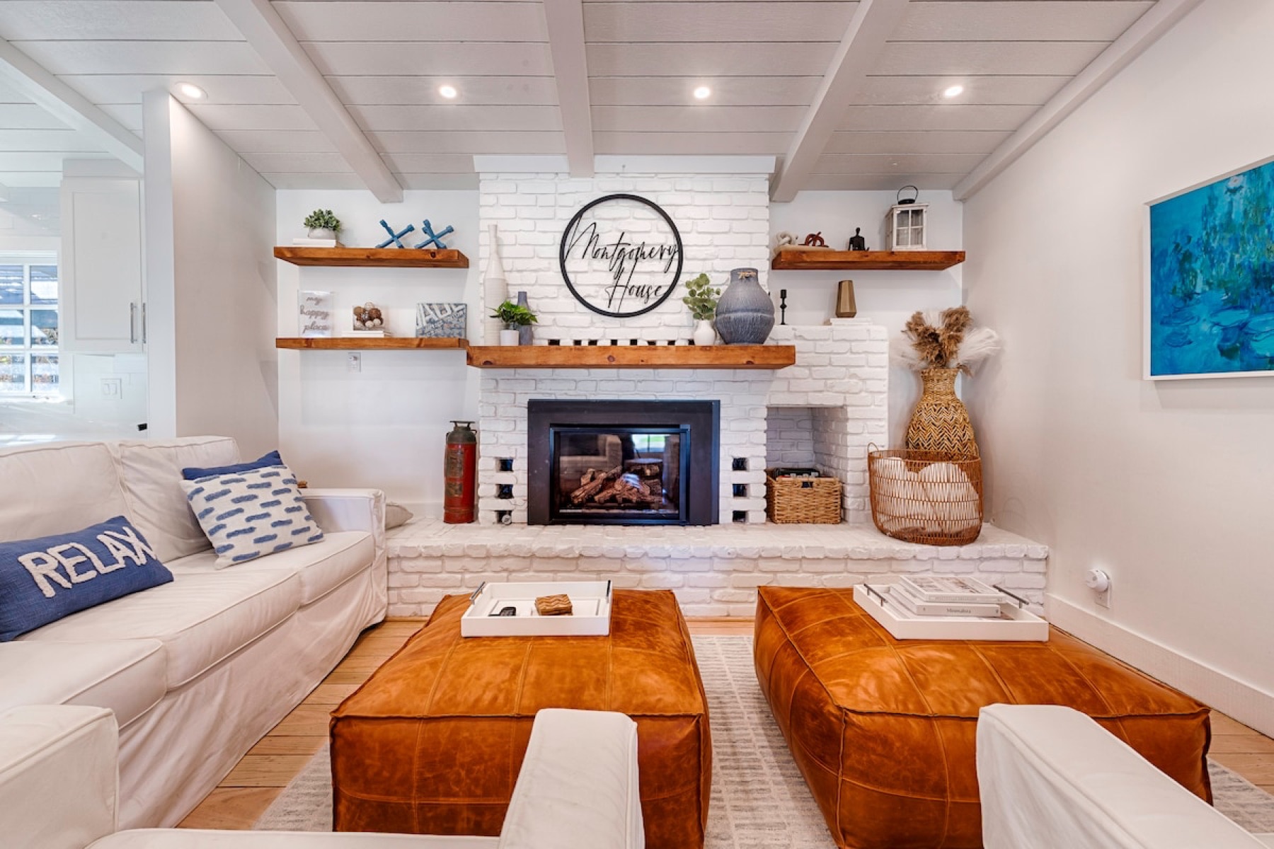 The long living room — a fire under whitewashed plank ceilings, leather ottomans, deep slipcovered sofas